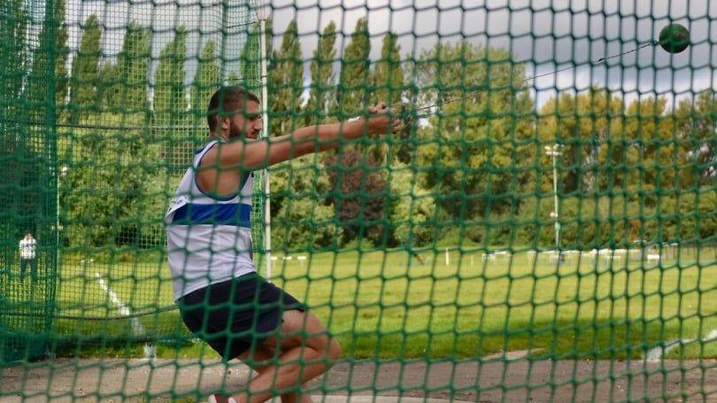 A man is using the sports facility and throwing a ball behind a net. The poplar trees can be seen in a row in the background. There is a playing field with markings between the man and the trees.