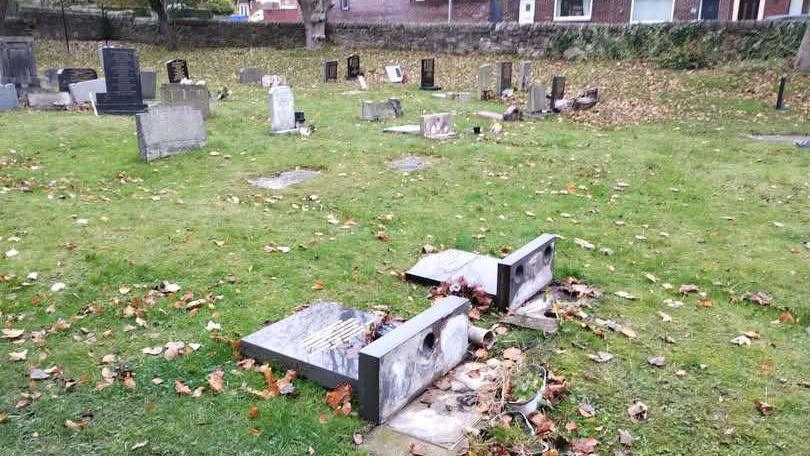 An open grassy cemetery with numerous headstones, some standing and others lying flat. In the foreground, two large gravestones are broken and toppled over, exposing their bases. Fallen leaves are scattered across the grass. A stone wall and residential houses can be seen in the background beyond the cemetery.