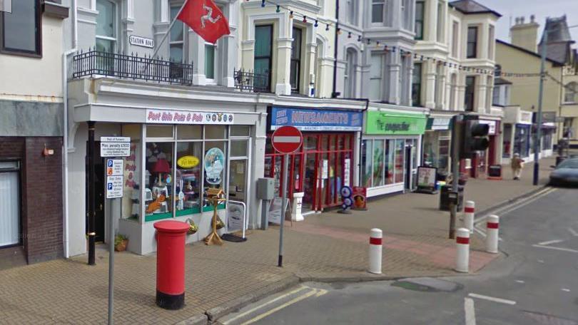 A row of shops on Station Road, including the Co-Op store. There are residential premises above the shops and paving in front. The road has a series of bollards around a traffic lights for a pedestrian crossing and there is a red post box to the left.