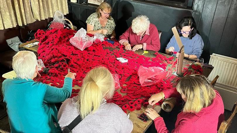 Another group of six women sit around a table, stitching the poppies together