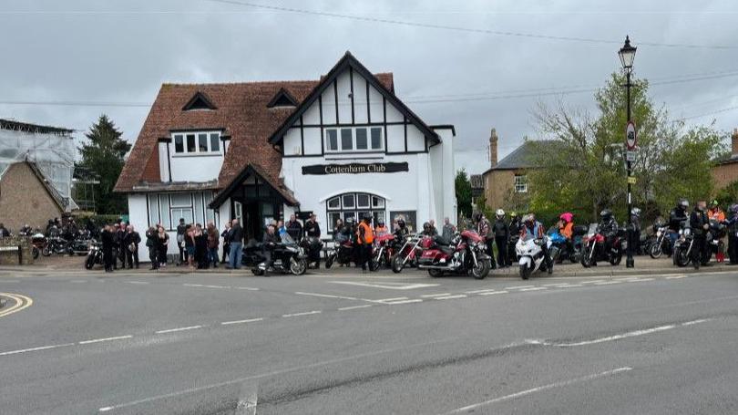 Lots of people in leathers with motorcycles are outside a club in Cottenham. The club is an old white building with timber frames