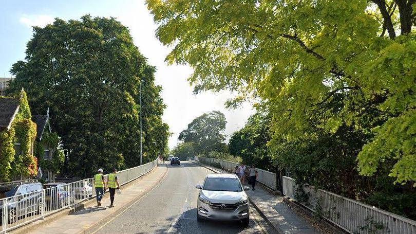 Two men in bright high visibility jackets walk along a bridge. people and a car are on the opposite side of the road. There are trees on either side of the road.