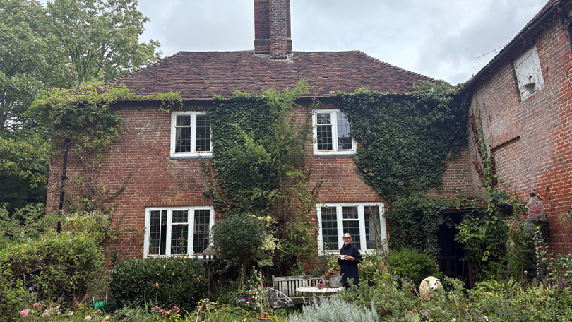 Baschea Walsh stands holding a mug in front of a cottage. It has two floors and four windows, parts of the front of the building are covered in ivy. To the right of Baschea, there is a life-sized model of a sheep.