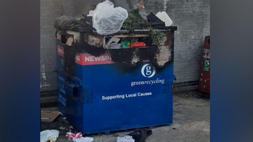 A blue recycling bin with rubbish pouring out of it. It is also burnt at the top so the blue bin has become discoloured and turned black.
