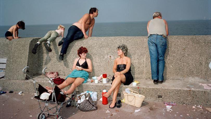 A family sitting on a stone wall with the sea behind them, they are enjoying a picnic and there is litter surrounding them on the floor 