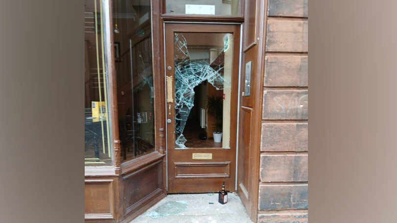 A brown framed door with glass in the centre that has been smashed. Broken glass lies on the concrete step outside the door with a bottle next to it.