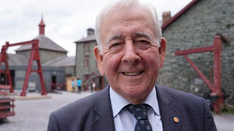 Smiling at the camera, a headshot of a white haired man, wearing a dark suit, blue shirt and navy tie, with a Plaid Cymru badge