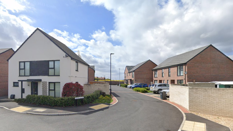 A photo of a short residential street, from the main road. A large white house with black window-frames and door is on the corner of the road, nearest the camera. Around four semi-detached brick homes are visible on the right hand side of the road. They look as though they are on a new-build estate, as they are quite uniform and appear to have been recently built. At the end of the road is hedgerow, and it looks like there is a field beyond. Each house has a car on the driveway.