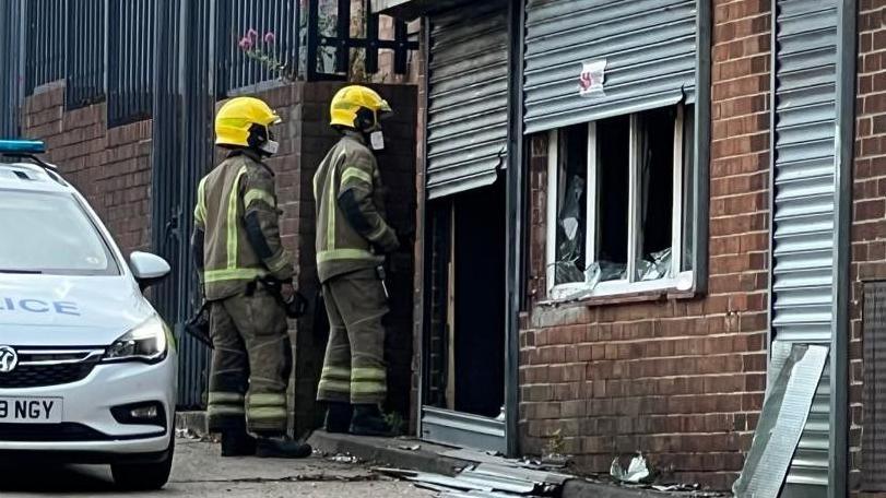 Two firefighters in uniform standing outside a fire-damaged brick building. The grey shutters are partially raised and the windows smashed. A parked police car can be seen outside the building, on the far left.
