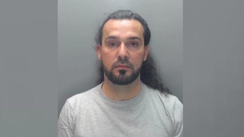 A police photograph of Ashkan Kareem. He wears a grey t-shirt and has long curly hair tied in a ponytail and a beard.