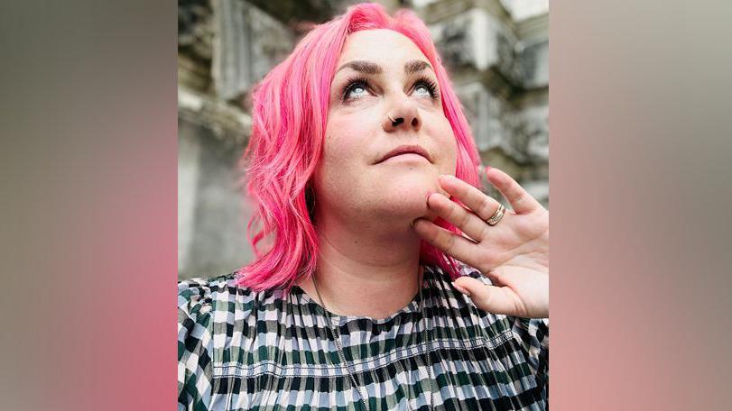 Emma with pink hair looking up with her hand under her chin. She is wearing a black top.
