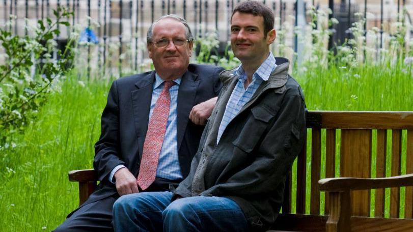 Two men, Donald MacDonald and Euan MacDonald sit on a bench. The man on the left has glasses and is wearing a navy suit, blue shirt and a pink tie. The man on the right is wearing jeans, a denim jacket and a blue checked shirt.