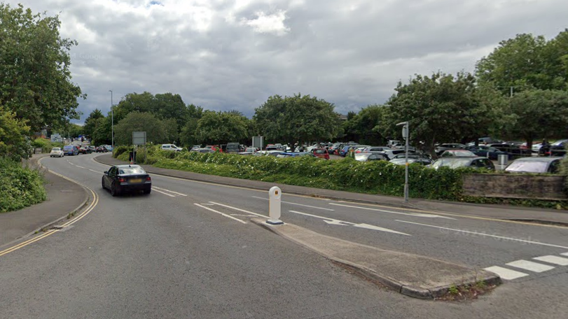 A Google Street View image of Bythesea Road. It is a wide main road, with a large car park on one side, and is bordered by green trees and shrubs