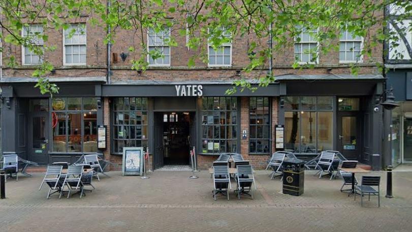 The outside of a high street bar with the word Yates over the door. Small tables and chairs are the front entrance. Branches of trees and leaves are overhanging the area. 