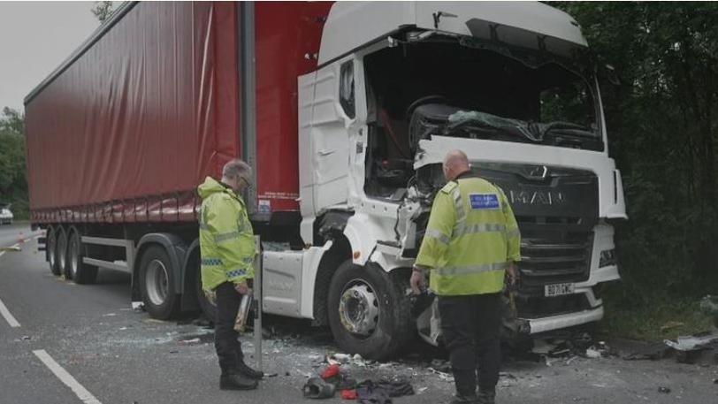 Two police officers look at crash wreckage. They are both in hi-vis. There is a red and white lorry with visible damage from a crash.
