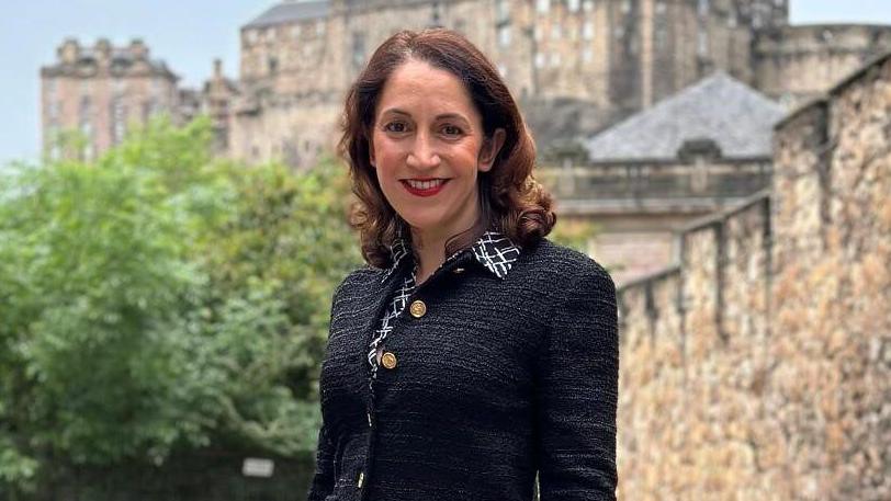 A woman with shoulder-length brown hair smiles while standing in front of Edinburgh Castle. She wears a black and white shirt and a black jacket