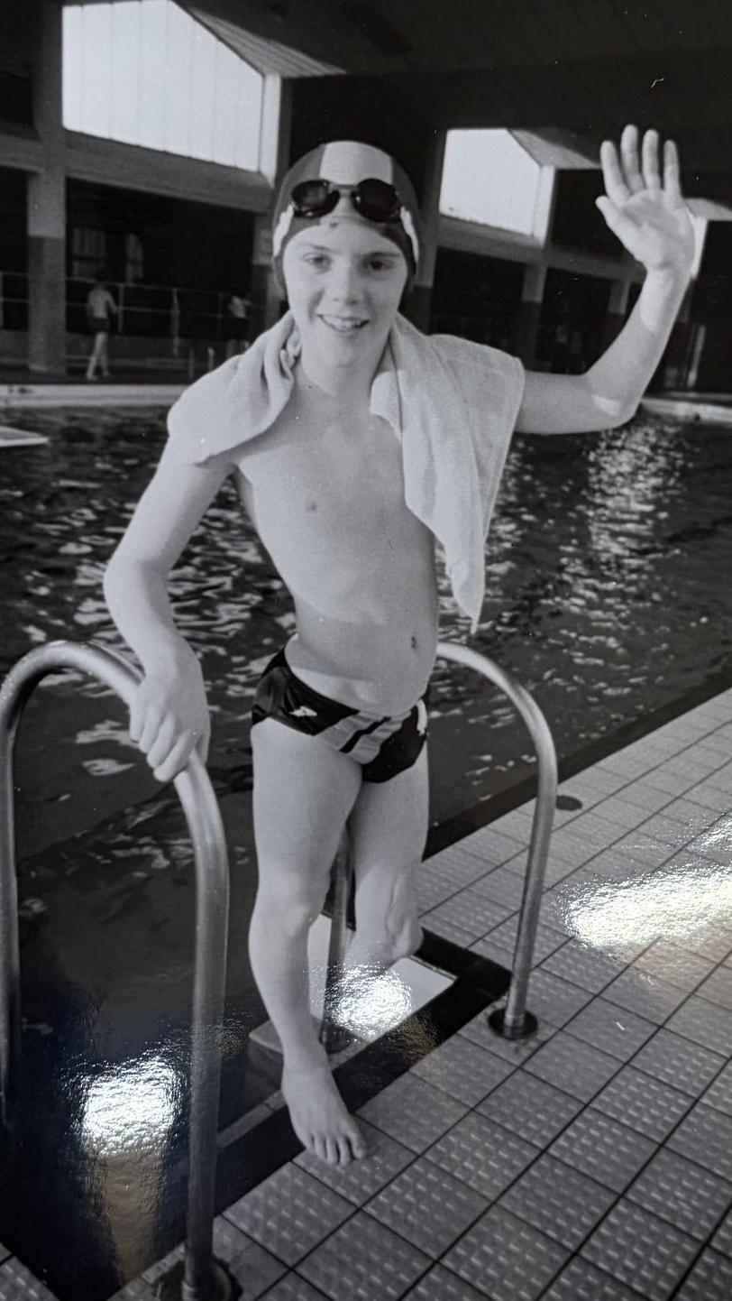 A black and white photo of Mark as a child, waving at the camera from the steps of a swimming pool. He is wearing swimming trunks and a swimming cap, and his left leg is amputated below the knee.