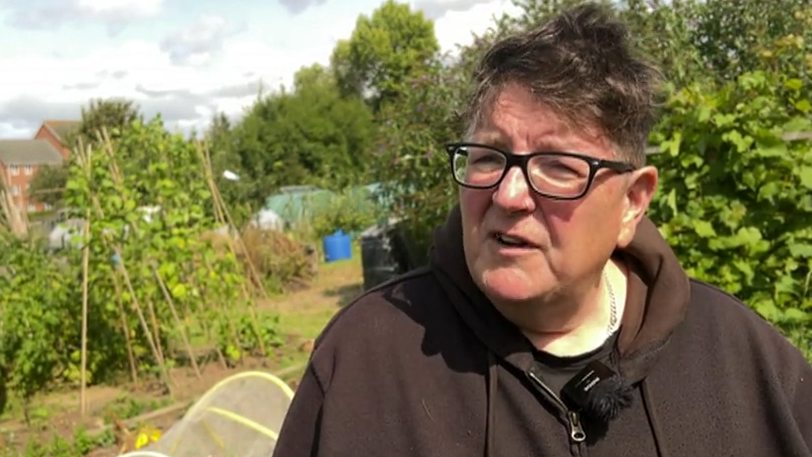 Sally Wyatt stood in her allotment. She is wearing a black jumper and glasses.