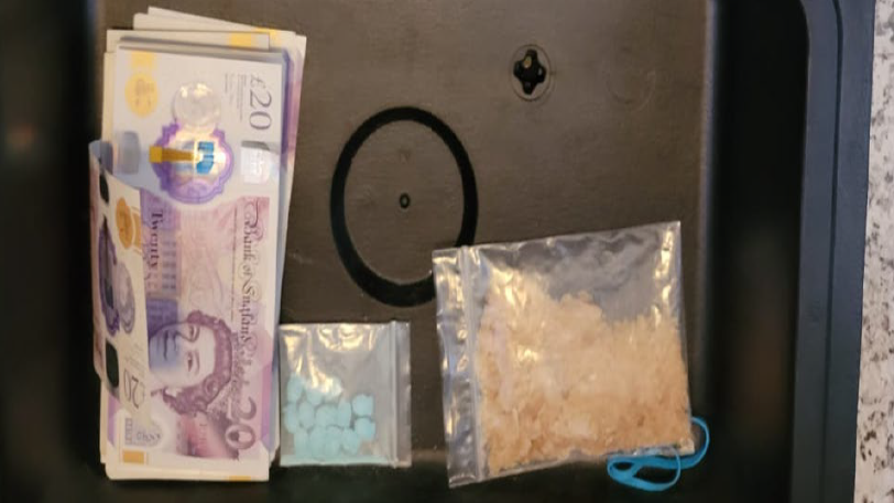 A stack of £20 notes in a black box next to two small plastic bags, one containing blue pills and another containing an orange substance.