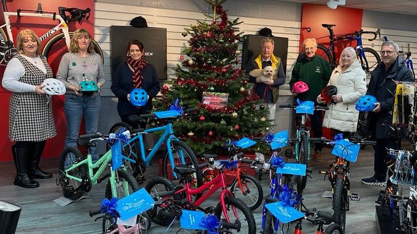 Seven adults holding bike helmets stood next to a Christmas tree.
They are in in front of a number of bikes with signs on saying Sarah Groves foundation