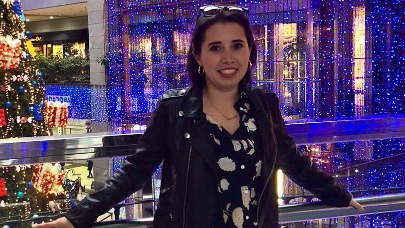 Marta Elena Vento poses on a balcony in a retail centre with Christmas decorations in the background. She wears a black leather jacket, a patterned black and white top and earrings and her glasses are pushed back over the top of her head.