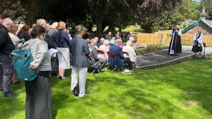 A shot of the backs of a crowd at the service outside under a tree. There are two priests in robes. Some people are sitting and their heads are bowed.