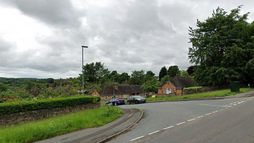 A road junction, with cars parked on the street which is dipping down a hill. There are street lights and houses as well as a pavement and a large tree near the junction.