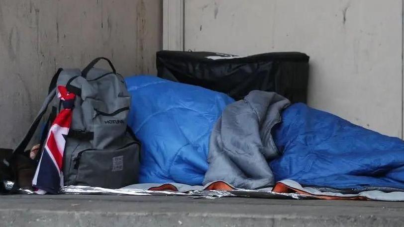 A person lies on the ground in a blue sleeping bag, rough sleeping. There is a grey backpack next to the person. There is a Union Jack flag tied to the bag.
