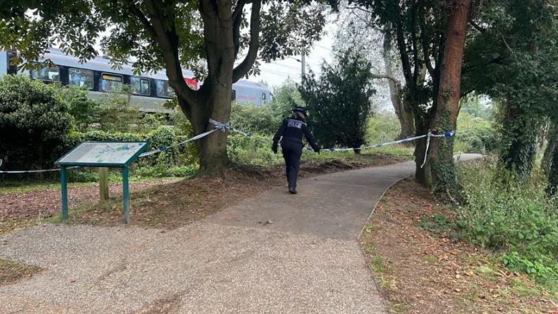 Police cordon put up around the lake with a police officer walking across the scene. There is a train coming past and lots of greenery in the photo with a footpath.