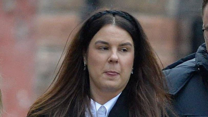 A woman with long brown hair wearing a white shirt and a black hairband looks away from the camera. 