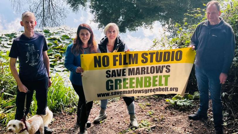 Four people stood near a body of water with Joy Morrissey (second left) holding a mostly yellow banner reading: NO FILM STUDIO. SAVE MARLOW'S GREENBELT". One man is holding a small white dog's lead, with the dog at his feet.