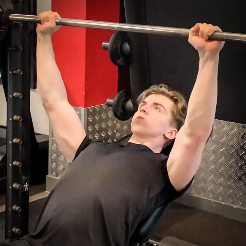 George Hazard a boy in a black t-shirt is pushing up a barbell weight while laying on a bench, carrying out bench presses