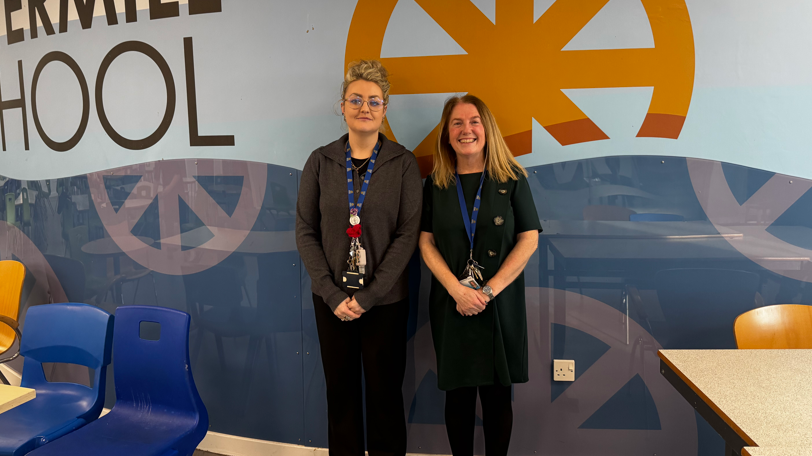 Two women stand in front of a large image on a wall, where the word school can be seen, as well as a repeated logo. Both have their hands crossed in front of them and are smiling.