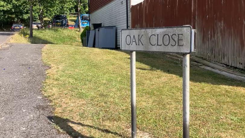 An image of a street sign riding Oak Close in front of a patch of grass and a footpath. A fence and some garages are visible in the background.