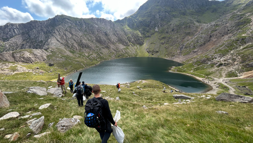 A group of people hiking down a grassy verge with a large body of water in front of them and mountains behind. They all have their backs to the camera and are holding rubbish bags and litter pickers.