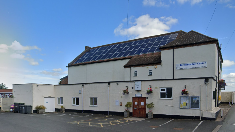 A white building with a sign that reads "Birchmeadow Centre". It has wooden double doors and solar panels on the roof, with a blue sky behind it