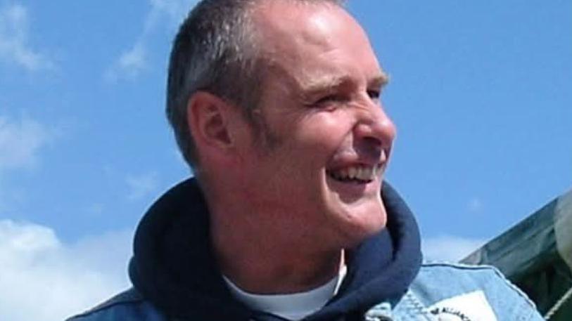 A photograph of Brian Rudkin, a middle-aged white man, wearing a denim jacket with a dark blue hoody underneath. He looking to his left and smiling.