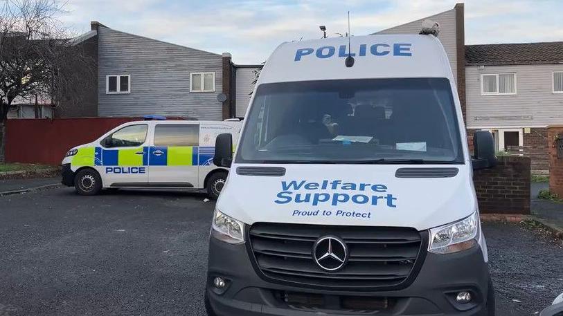 A grey police welfare support van parked in the housing estate in Elswick. Behind it and to the left, another police van is parked. A row of terraced houses stands behind the vans.