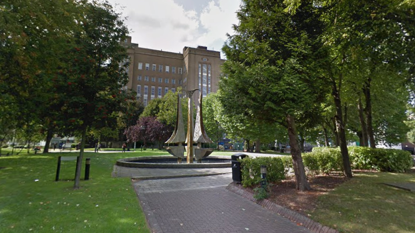 An early 20th-Century brick building, approximately six stories high, is flanked by trees and green space with a modern sculpture, Tipping Triangles, in a water feature in the foreground. The sculpture consists of three 18-foot stainless steel "triangles" with one curved side.