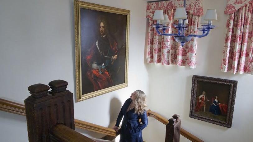 A woman wearing a long blue dress stares up at a painting as she walks up a grand wooden staircase. The painting appears to be a portrait of a nobleman from the 17th or 18th Century. On another wall is a painting with two curtains above in a pink and white toile pattern