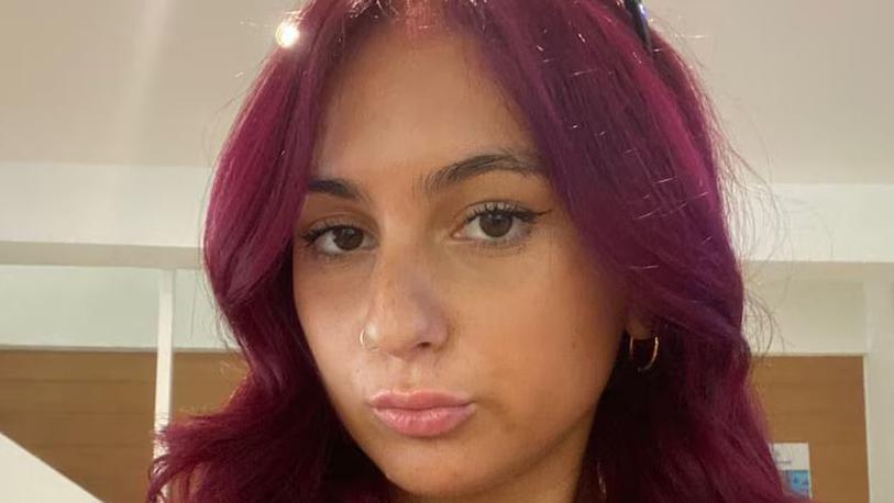 Alicia Kemp, looking at camera, with a pout. She has long hair dyed a reddish purple colour. 