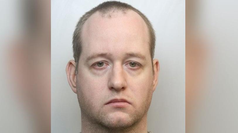 Mugshot of a man wearing a grey shirt. He has very short hair and stubble.