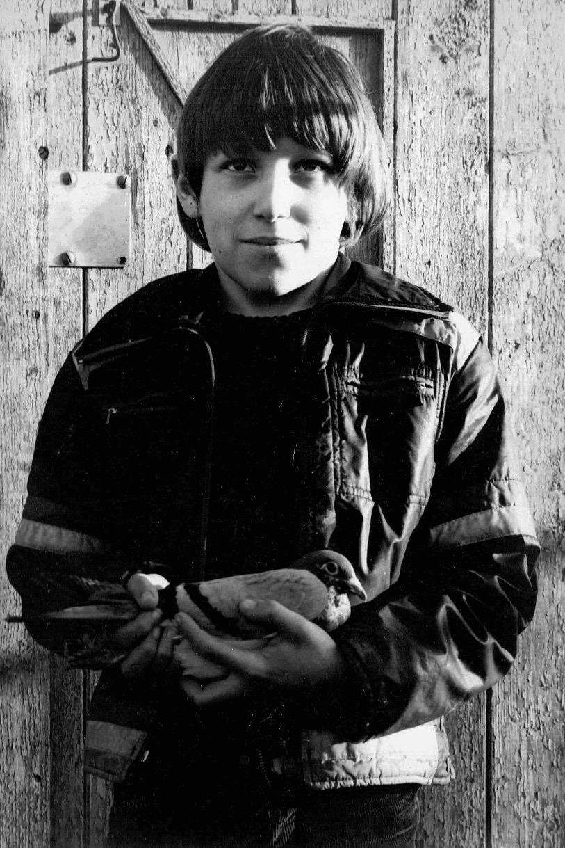 A black-and-white photo of a young boy holding a pigeon in his arms. He has short, floppy hair and is looking above the camera with a slight smile. He is wearing dark clothes. The pigeon looks settled in his arms.