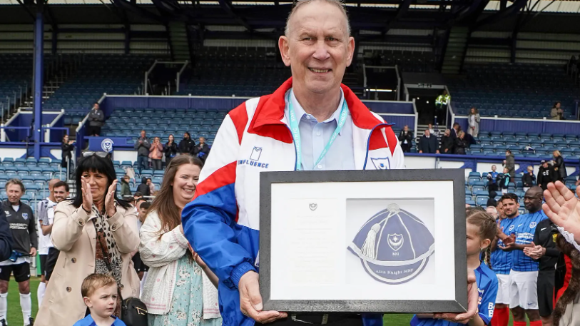 A man with very short hair and wearing glasses on top of his head holding up a framed honour. He's wearing a blue, red and white jacket and smiling at the camera. Behind him, crowds of people are clapping in front of blue raked seating.