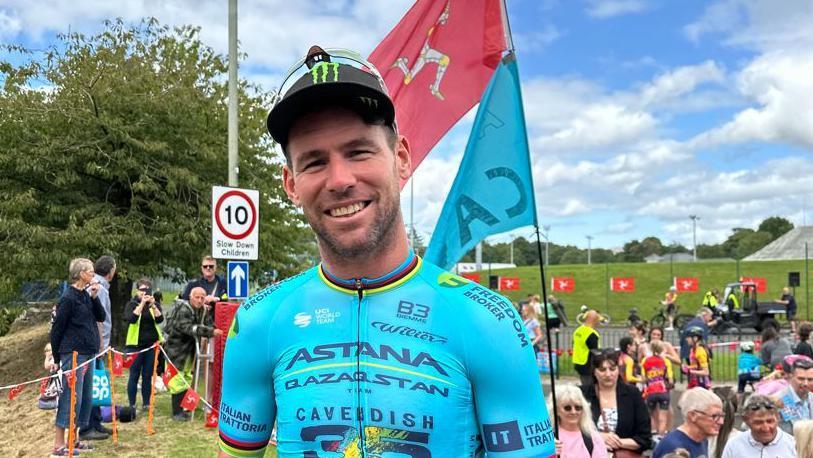 Sir Mark Cavendish in his blue 35 stage victory jersey at the sports centre raceway. A Manx flag is behind him with crowds.