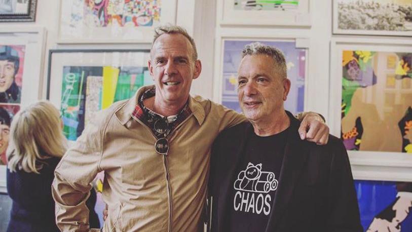 Fatboy Slim with his arm around Lawrence Alkin, who is wearing a black t-shirt and a black jacket. Fatboy Slim is wearing a cream jacket. 