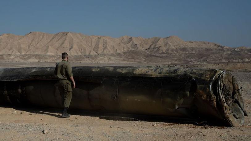 An Israeli soldier inspects remains of a missile launched by Iran into Israel on 1 October. The large missile is lying in the desert, and the official stands in front of it with his back to the camera. 