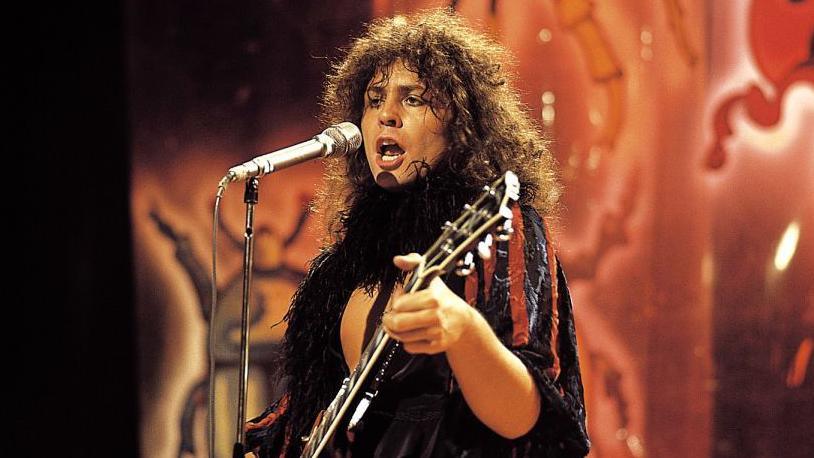 An image of Marc Bolan, a man with long curly hair playing a guitar and singing into a microphone, performing wearing a blue and black velvet jacket and black feather boa. Behind him a background painted with colourful beetles can be seen