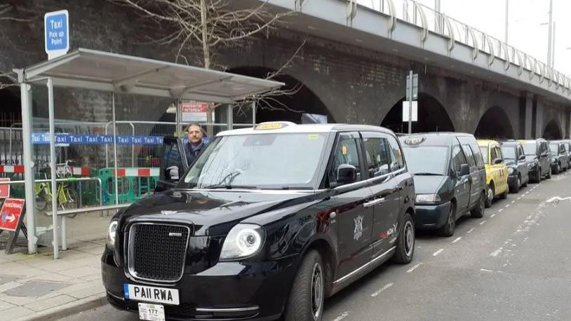 A man at the door of his black cab in the taxi rank, a line of taxis is in view behind it. There is a shelter with a sign that says "taxi rank pick-up point"