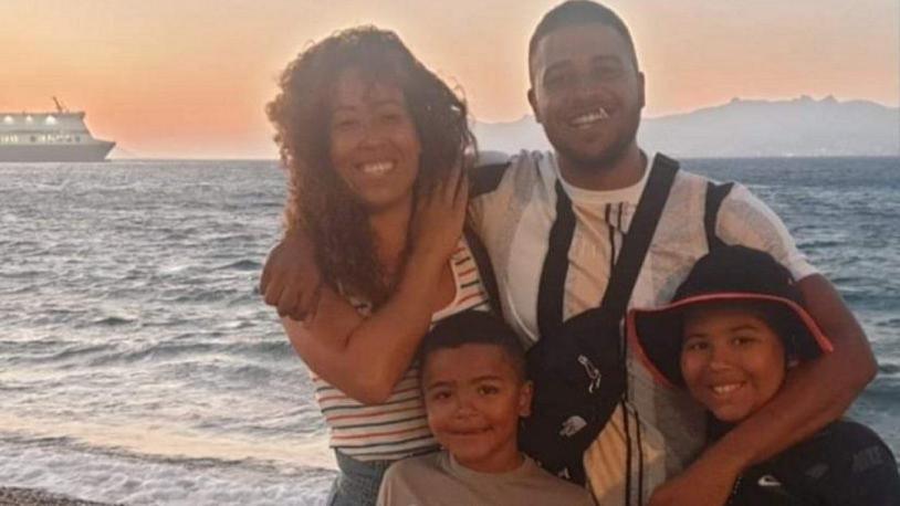 Lorette Divers is pictured with her partner and two children at a beach during a sunset. The ocean can be seen in the background as well as a large vessel in the left corner. Some mountains or cliffs can be seen on the right. Ms Divers has curly brown hair and is smiling at the camera. Her partner has his arm around her and one of their sons, who is wearing a hat.